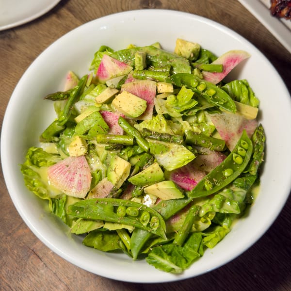 A vibrant green salad with lettuce, avocado, asparagus, snap peas, and watermelon radish in a white bowl.