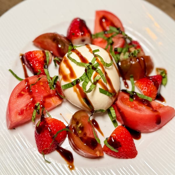 A burrata cheese ball with basil, surrounded by sliced tomatoes and strawberries, drizzled with balsamic glaze.
