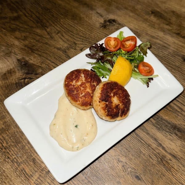 Two golden-brown crab cakes with creamy sauce, lemon wedge, and a side salad on a white plate.