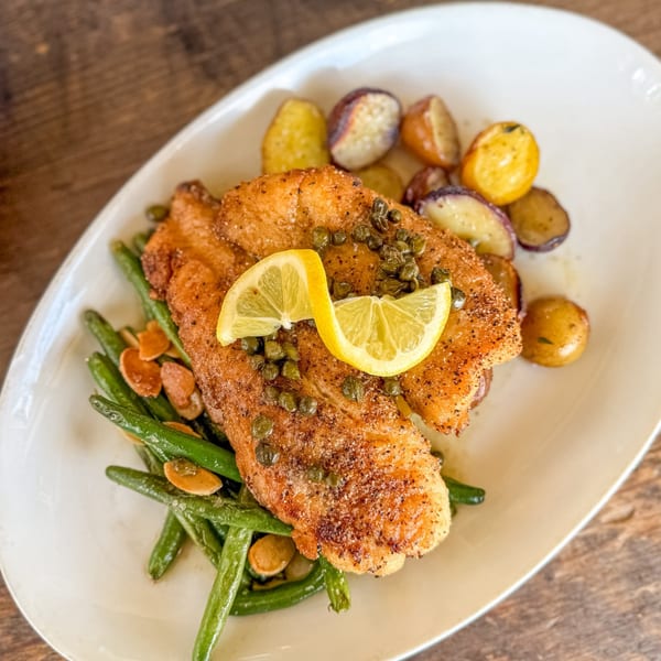 Pan-seared fish with capers, lemon, green beans, almonds, and roasted potatoes on a white plate.